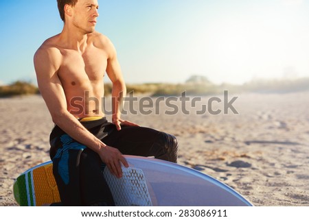 Topless Athletic Young Man Sitting on the Edge of his Surfboard at the Beach Sand on a Tropical Climate While Looking Into Distance.