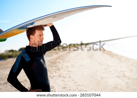 Attractive surfer balancing his surfboard on his head as he stands on a sandy tropical beach looking out over the ocean and surf, close up profile view