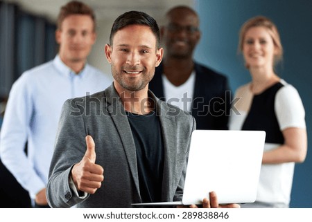 Young white executive making thumbs up sign holding laptop facing camera with colleagues in background