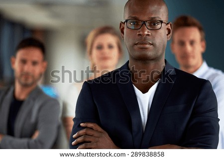 Black male executive with arms crossed and serious expression facing camera with colleagues in background