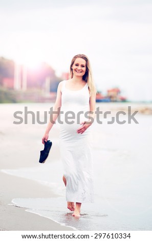 Happy pregnant woman at the seaside walking along barefoot at the edge of the surf in a white maternity dress smiling at the camera