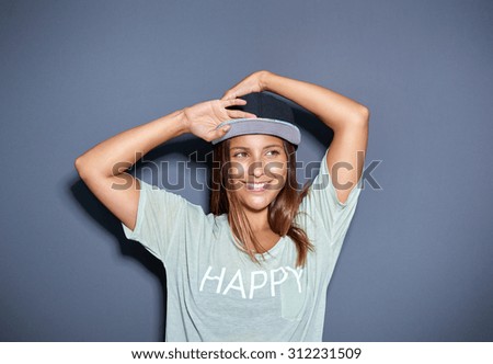 Pretty trendy young Asian student with a lovely friendly smile posing with her arms raised above her head in a baseball cap and t-shirt over a grey background, looking to the right