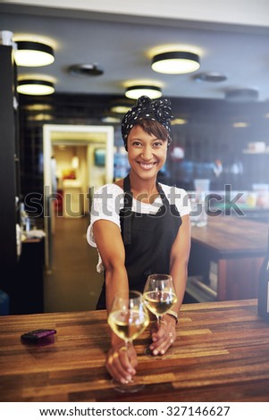Pretty successful African American businesswoman standing behind the wooden counter in her pub serving glasses of white wine to a customer with a warm friendly smile, small business owner