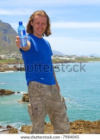 Smiling man offers water bottle standing on beach among awesome mountains. Shot on Cliff Path near Hermanus, Walker Bay, Western Cape, South Africa.
