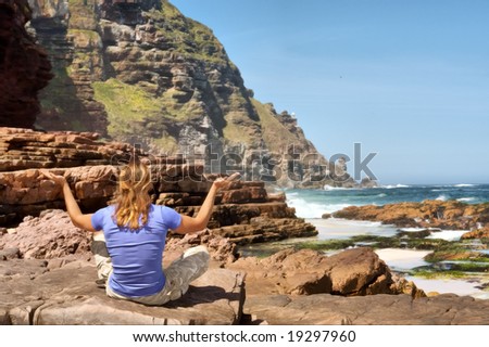 Blond young man meditates on rocks against sea-misty mountain background. Shot in Cape of Good Hope n Cape Point Nature Reserve, Table Mountain National Park, near Cape Town, South Africa.
