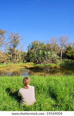 Blonde woman sits next to small pond in park. Shot in Vergelegen estate, near Cape Town, Western Cape, South Africa.