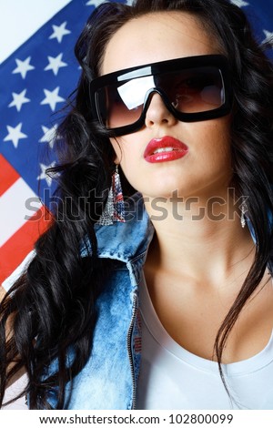 beautiful young brunette woman in style cloth and sunglasses with national usa flag in background with copyspace. 4th july concept