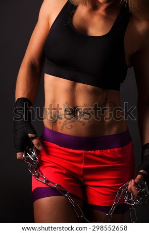 A very muscular girl is holding a chain in her hands. Detail shot of belly muscles with side light.\
The girl is tattooed.