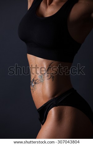 Wet body of a muscular women with a tattooed belly. Side light picture. Women hold the hand in front of the breasts.