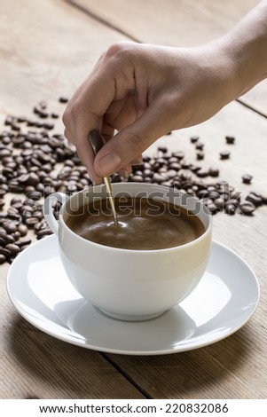 Image of a coffee cup being stirred by a white human hand, on a wooden table top surrounded by raw coffee beans