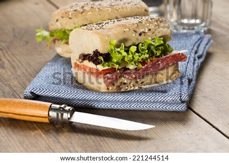 Image of a sandwich with red tomato, fresh lettuce and cheese, covered with sesame seeds wrapped in wrapping paper, on a blue napkin with a sharp knife