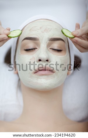 Beautiful young woman receiving facial mask of cucumber in beauty salon, hands of cosmetologist