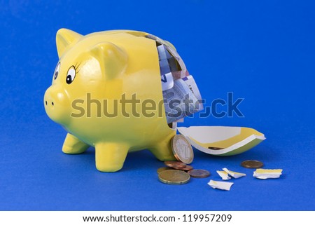 Yellow broken piggy bank containing euro bills and coins on blue background