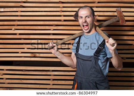 Angry worker with axe and hammer. Horizontal shot with wood in the background.