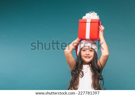 Smiling funny child (kid, girl) in Santa red hat. Holding Christmas gift in hand. Christmas concept. Shooting on blue background