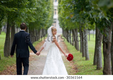Just married couple is walking down the path in the park. The bride a wedding bouquet of red roses is turning back and smiling to the camera