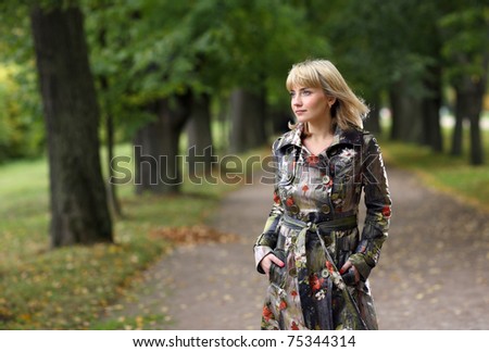 The fair-haired woman in an autumn coat. Walk in autumn park. The fallen down leaves on a path, the big trees.