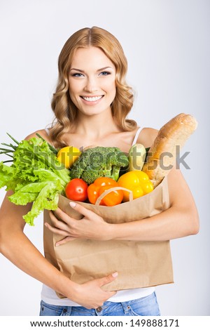 Portrait of happy smiling young beautiful woman holding grocery shopping bag with healthy vegetarian raw food, over gray background