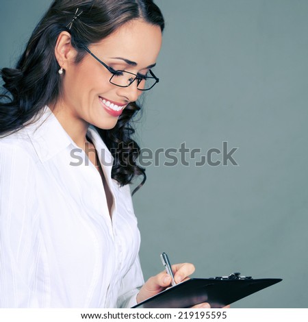 Happy smiling young beautiful business woman with clipboard writing, over gray background