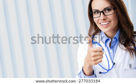 Happy smiling young female doctor in glasses, showing thumbs up hand sign gesture, at office, with blank copyspace area for slogan or text