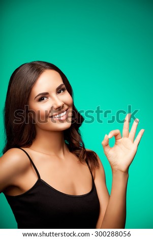 Portrait of young smiling woman in black tank top clothing, showing okay gesture, with blank copyspace area for text or slogan, on green background