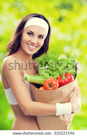 Happy smiling young lovely brunette woman in fitness wear holding grocery shopping bag with healthy vegetarian raw food, outdoors. Healthy lifestyle, beauty and dieting concept.