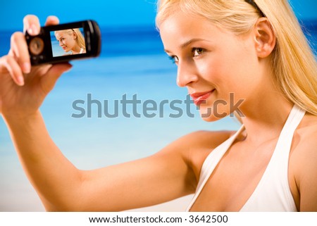 Young beautiful tanned happy smiling blond woman in bikini taking photograph by cellphone on beach