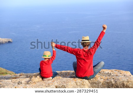 father and son sitting on top of a mountain expressing joy with their arms stretched up towards sky