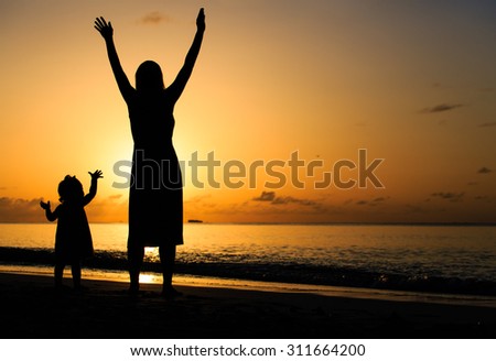 silhouette of happy mother and little daughter hands up at sunset beach