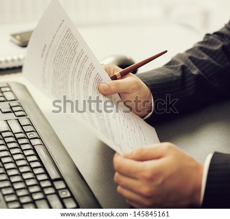 Close-up of company director\'s hands holding business document at the computer desk. Young financial specialist working over calculation papers.