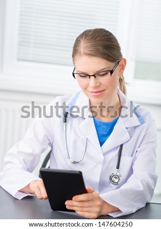 Nice woman doctor sitting at the desk and pointing on tablet PC. Beautiful blond Caucasian medical female with stethoscope, ipad wearing eyeglasses in hospital.