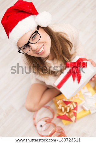Beautiful girl in red Santa hat and eyeglasses holding xmas gift sitting on the floor at home in cold winter weather. Nice blonde wrapping Christmas gift boxes.