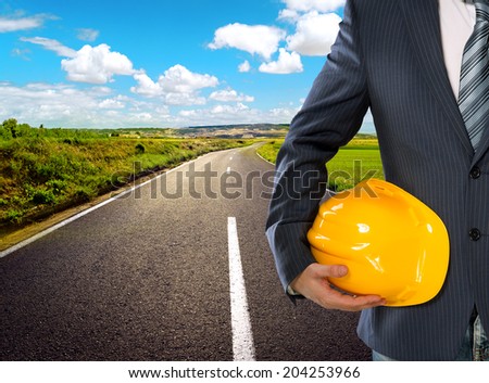 Male engineer on rural road through agricultural meadow. Business man holding yellow hardhat for safety of the workers on sunny landscape background.