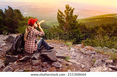 Woman climber with binocular looking at green hills landscape. Young active female traveler sitting on stones on top of the mountain.