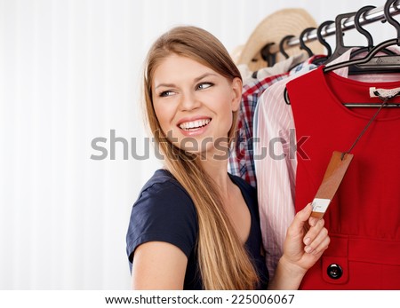 Young female shopper holding price tag of new dress in retail store. Portrait of attractive joyful shopping woman dreaming of buying clothes.