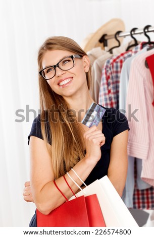 Cheerful young lady with shopping bags and plastic card buying clothes in retail store. Happy female customer posing at the rack with clothing.