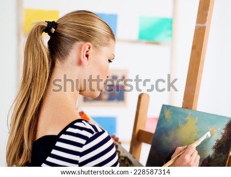 Woman painter creating new artwork in her artistic studio. Young female artist drawing oil picture of landscape.