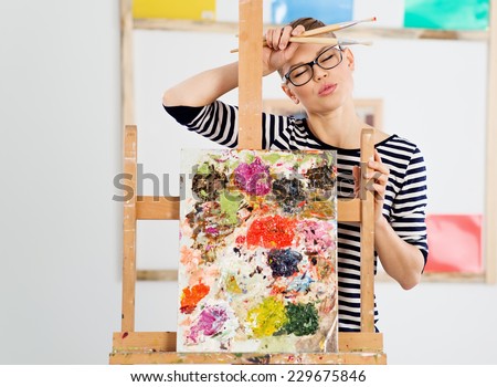 Tired woman artist standing with brushes at canvas in studio. Portrait of female painter weary after working on picture.