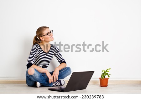 Young smiling woman dreaming of new home decor and furniture. Pretty Caucasian female sitting with laptop at white wall in empty room of new bought apartment.