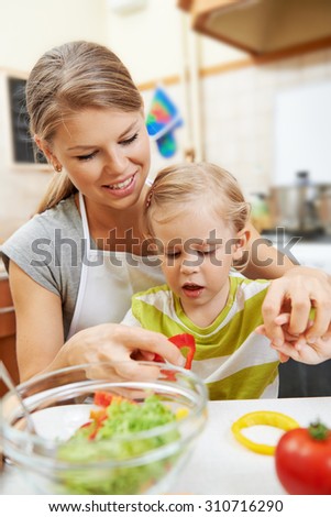 Lovely baby girl helping mother with meal in the kitchen. Young smiling female with little daughter cooking vegetables for family dinner.