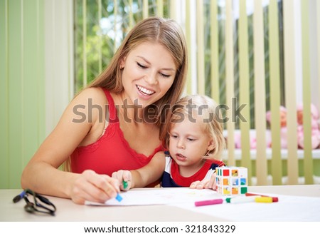 Smiling female and her small daughter drawing with colorful chalks at the table. Concept of child and preschooler development.