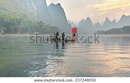 XINGPING, CHINA - OCTOBER 22, 2014: Fisherman stands on traditional bamboo boats at sunrise (boat with a red sail in the background) - Li River, Xingping, China