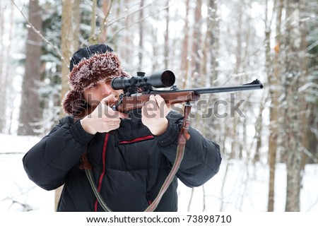 Hunter in a fur cap with ear flaps with sniper rifle in winter forest.