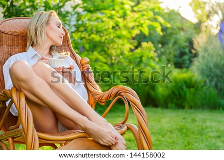 Young blonde woman wearing in man\'s shirt relax at outdoor sitting on a rocking chair holding tea cup