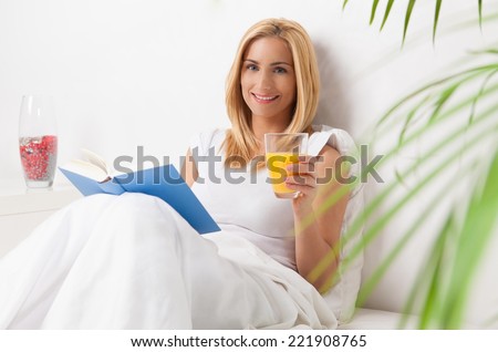 Young woman in bed reading a book and drinking orange juice.