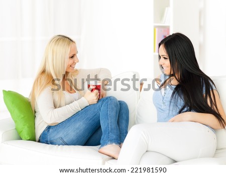 Two sisters sitting on the sofa at home and talking. Blonde and dark haired girl.