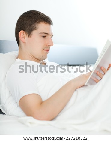 Young man in bed reading a book and drinking tea or coffee.