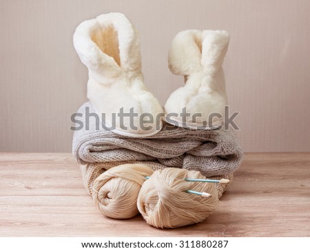 Pair of warm winter sheepskin slippers (alpaca), wool yarn , knitting needle ,  pile of warm woolen clothes (Selective focus)