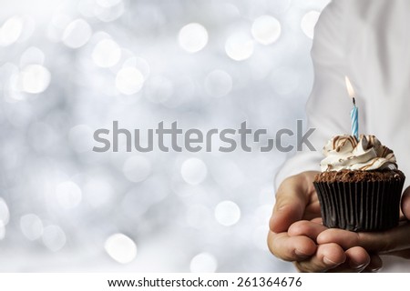 A Man holding a cupcake with a candle in a silver lights background.