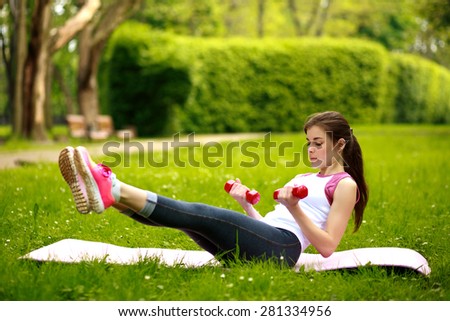 Sportive young woman stretching with dumbbells, doing fitness exercises in green park